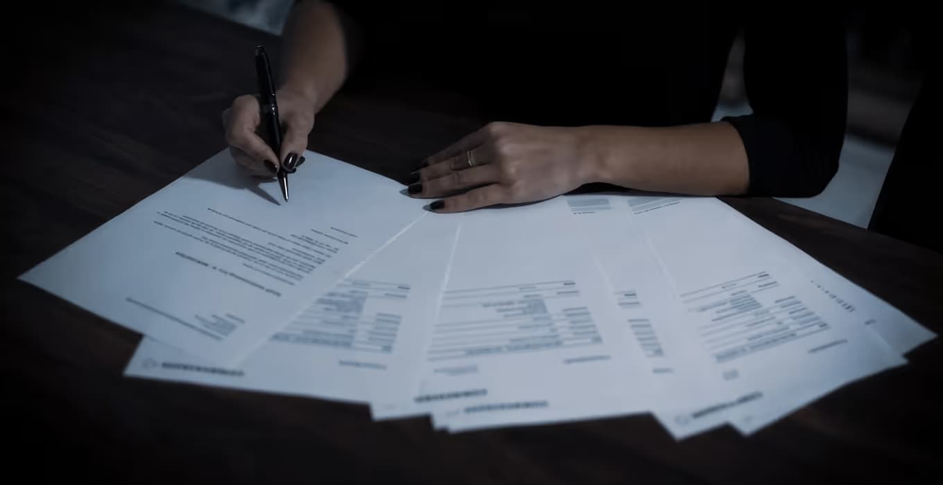 Hands signing a fan of contract documents with a pen on a dark wooden desk